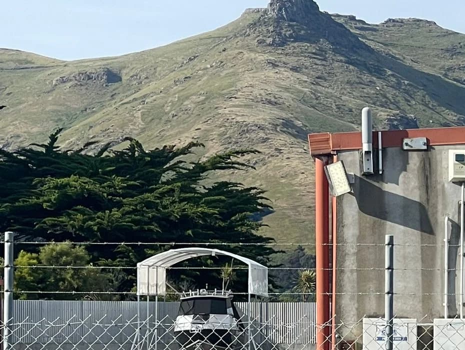 A boat stored under cover beside a concrete unit, with the Port Hills behind