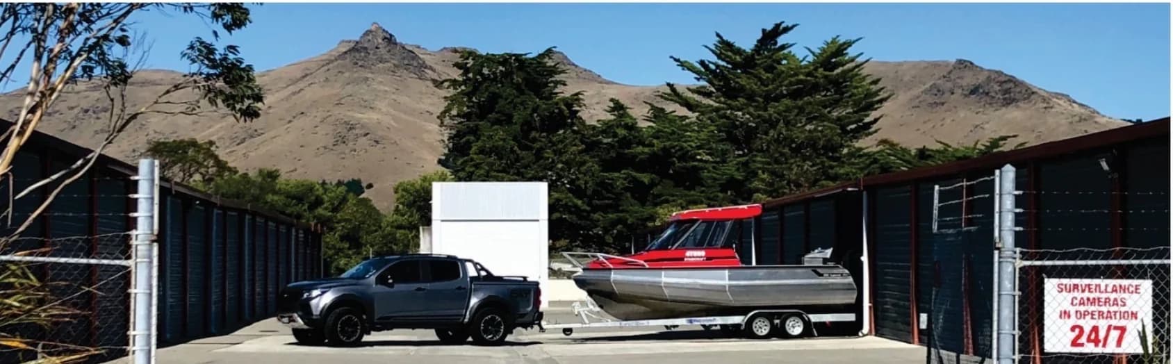 A ute towing a boat inside the Ferrymead Storage yard, with the units on both sides and the Port Hills behind